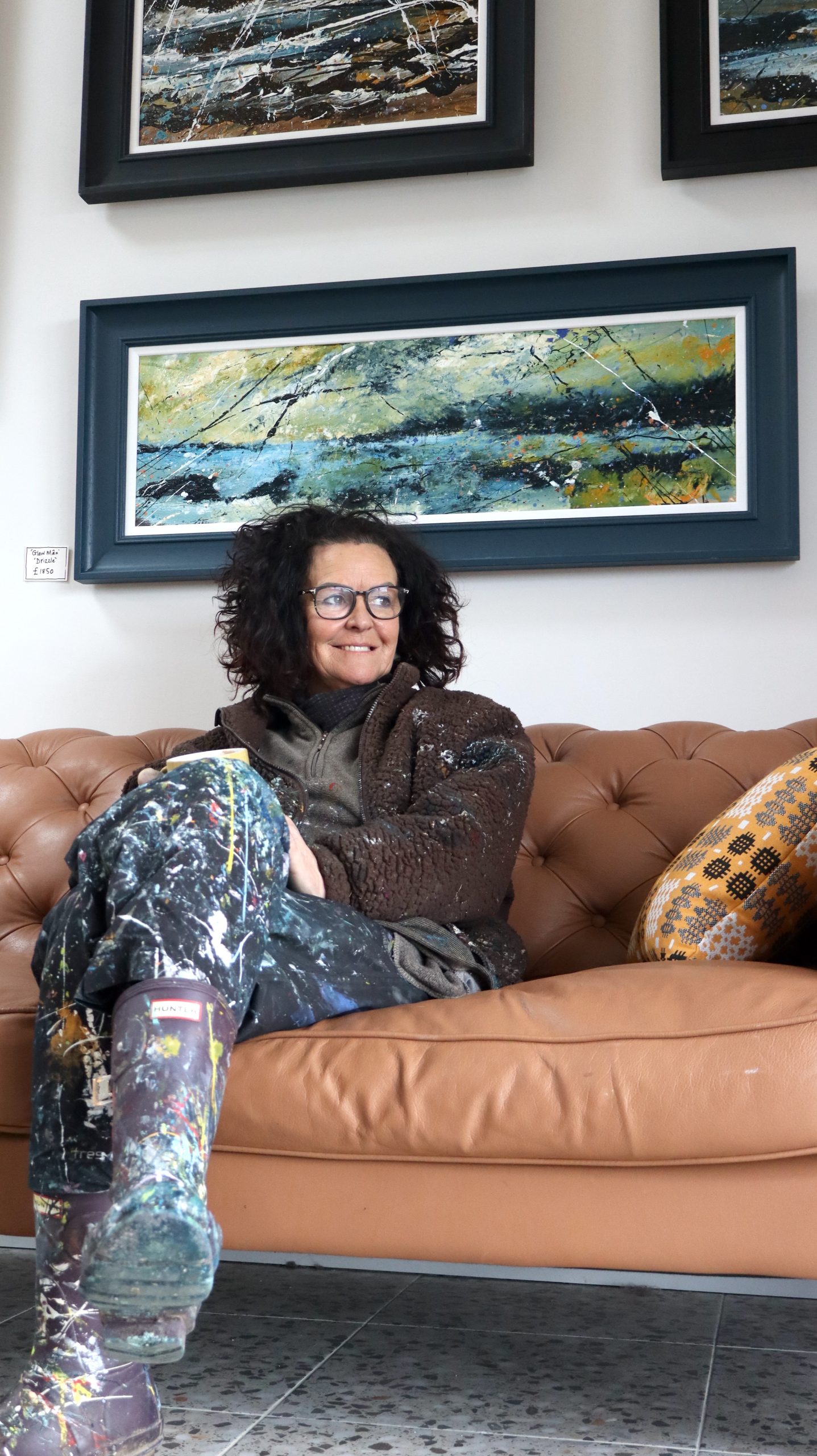 The artist Carys Bryn sitting on a sofa with a cup of tea and her paintings behind her on the wall.
